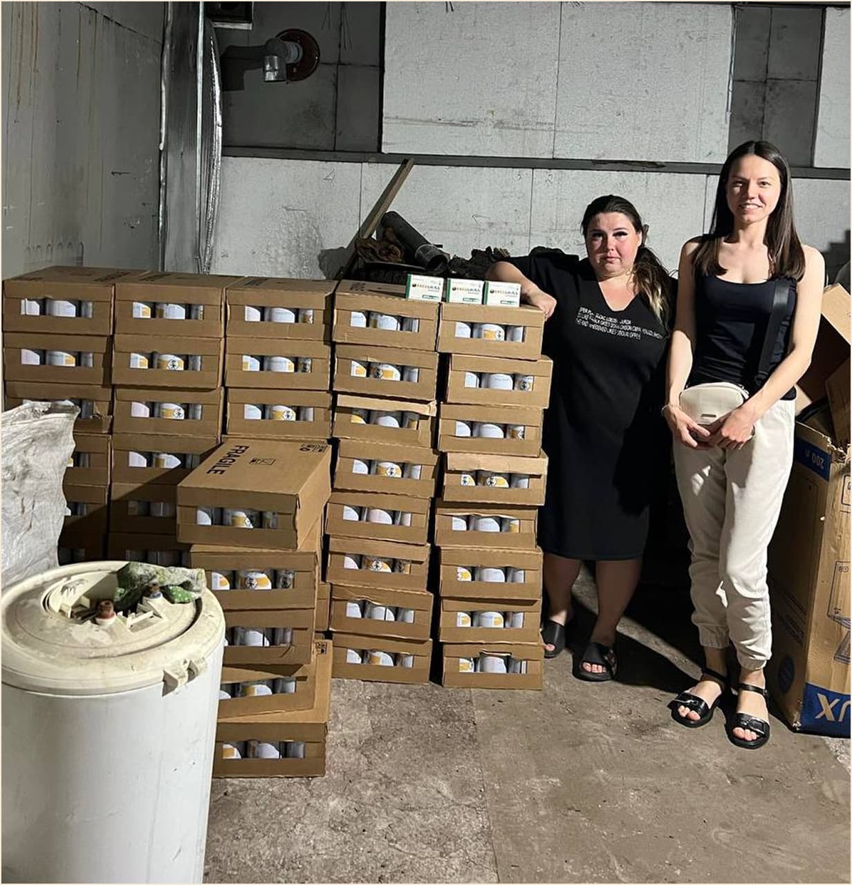 Two women standing next to stacks of boxed supplies in a warehouse-like setting, representing a donation or aid delivery